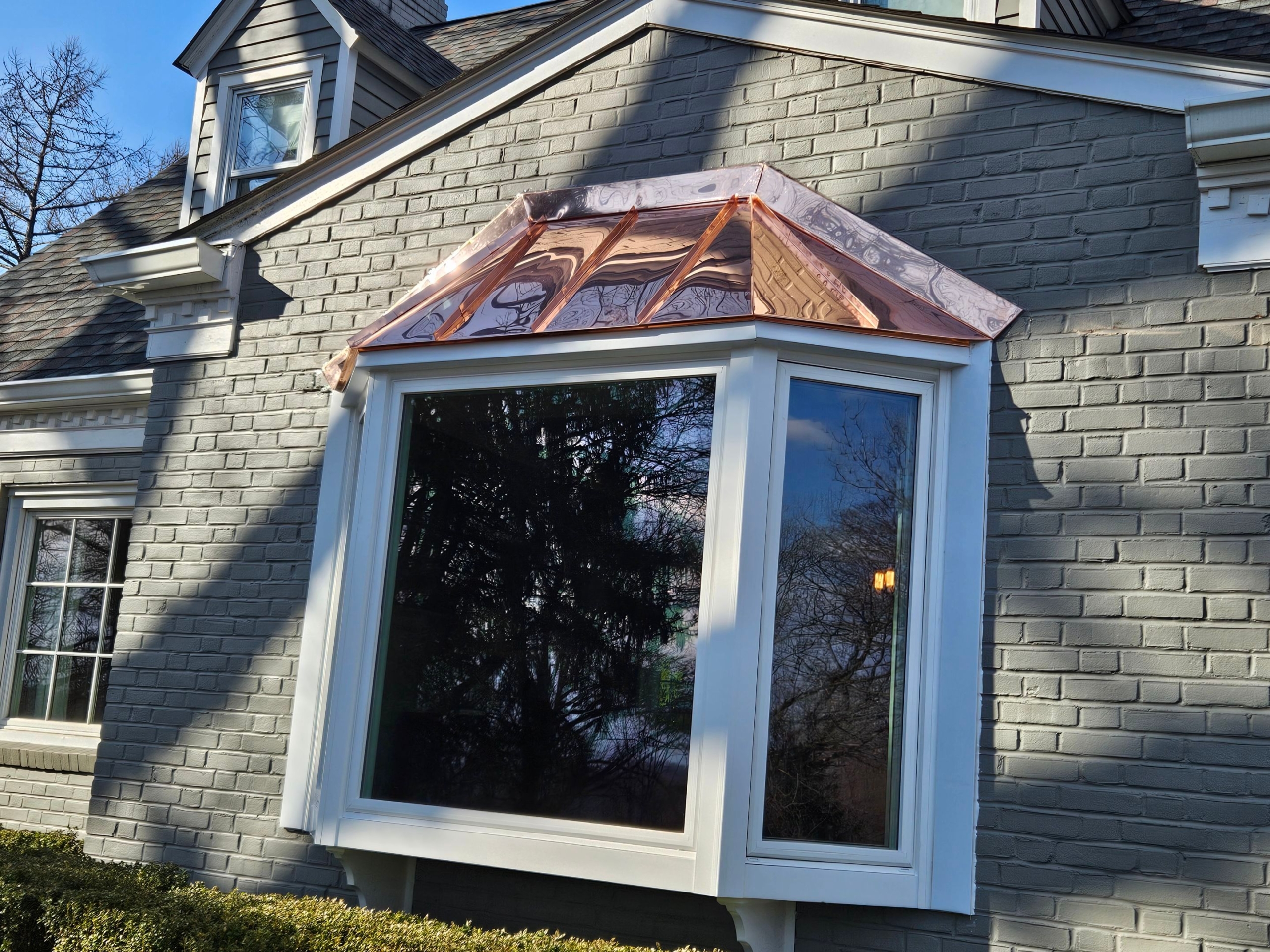Bay window with newly installed copper roof on a gray brick house exterior, featuring white trim and reflecting trees in the window glass.